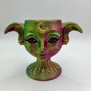 May include: A decorative, hand-painted planter with a face design. The planter features a green and purple colour scheme with gold accents. It has large ears, long eyelashes, and a crescent moon detail. The base is adorned with star and swirl patterns.