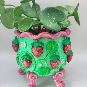 May include: A decorative planter pot, hand-painted in green, pink, and red, featuring strawberry and floral motifs. The pot has decorative legs and contains a leafy green plant. A charming piece for home decor.