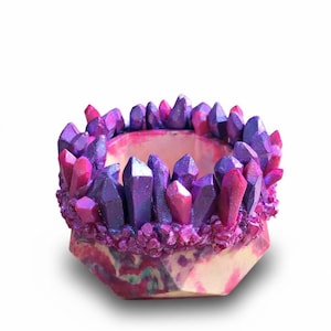 May include: A decorative, hexagonal container with a crystal design. The rim is adorned with purple and pink crystal formations. The base features a marbled pattern in shades of pink, purple, and cream.