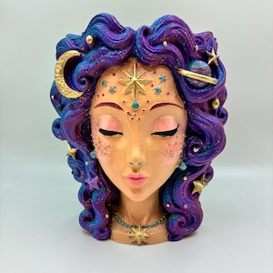 May include: A decorative head-shaped planter featuring wavy purple and blue hair adorned with gold stars, a crescent moon, and a small planet. The face has closed eyes, pink cheeks, and a gold star on the forehead, with a beaded necklace and star pendant.