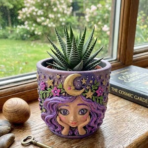 May include: A purple planter featuring a sculpted face with wavy purple hair, a gold crescent moon, and pink flowers, containing a green and white striped succulent plant. The planter has a textured surface and a detailed, hand-painted design.