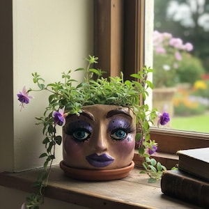 May include: A decorative ceramic face planter with a terracotta saucer. The planter features a painted face with blue eyes, long eyelashes, and purple lips. Green plants and purple flowers spill over the top, creating a unique and artistic display.