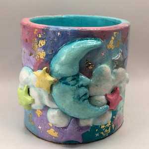 May include: A cylindrical ceramic container with a celestial theme. The exterior features a gradient of pink, purple, and blue with gold leaf accents. A turquoise crescent moon, stars, and clouds are sculpted on the front. The rim is pink and the interior is turquoise.