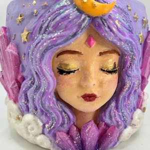 May include: A decorative planter shaped like a face with wavy purple hair, gold eyeshadow, and a yellow crescent moon on the forehead. The design features pink crystal-like protrusions on the sides and bottom, gold stars, and white clouds.