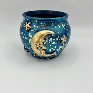 May include: A round, deep blue ceramic pot featuring a prominent gold crescent moon with a face, gold star accents, and scattered gold leaf flakes. The surface is decorated with small, iridescent rhinestones throughout the exterior.