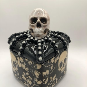 May include: A black ceramic box with a cream floral and skull pattern. The lid features a skull and is surrounded by black beads. The sides of the box also feature a skull design. The box is a decorative item.