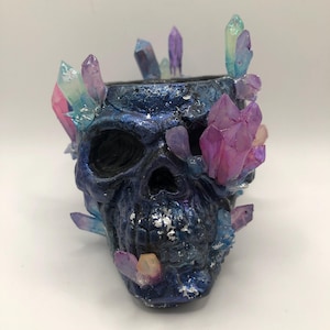 May include: A dark blue skull-shaped container decorated with colourful crystals. The crystals are various sizes and colours, including pink, purple, and blue. The container has a textured surface with silver accents.