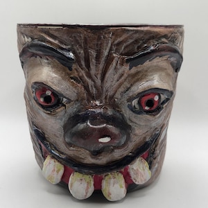 May include: A brown, hand-painted ceramic planter with a monster face design. The planter features red eyes, a black nose, and large, white teeth. The overall design has a textured, aged appearance.