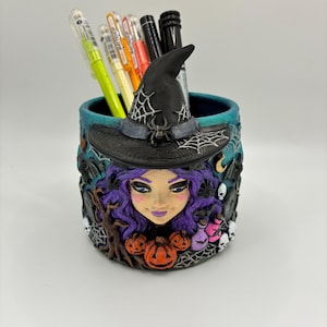 May include: A cylindrical teal pen holder featuring a sculpted witch with purple hair wearing a black hat with a spider and web design. The exterior is decorated with small orange pumpkins, white skulls, bats, and trees. Several pens are inside.