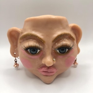 May include: A ceramic face planter with blue eyes, long eyelashes, and pink blush. The planter has a nose ring and earrings with pearl-like drops. The face is a light skin tone with a neutral expression.