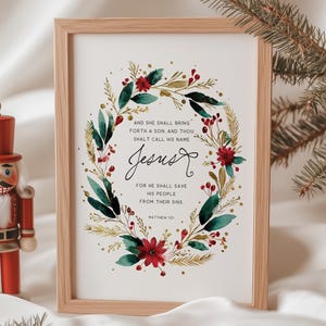 Peut inclure: Une impression encadrée avec le mot "Jésus" en écriture élégante, entourée d'une couronne de feuilles vertes, de fleurs rouges et d'accents dorés. Un casse-noisette rouge est à gauche. Le texte dit "Et elle enfantera un fils, et tu lui donneras le nom de Jésus."