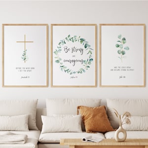 May include: Three framed prints with inspirational quotes and watercolor greenery. The first print features a gold cross with the text "Before you were born I set you apart Jeremiah 1:5". The second print features a wreath of green leaves with the text "Be strong and courageous Joshua 1:9". The third print features a sprig of green leaves with the text "And the child grew and became strong in spirit Luke 2:40".