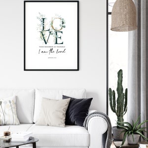 Op de afbeelding: Een zwart ingelijst print met een witte achtergrond waarop het woord "LOVE" in grote letters staat met groene en gouden bloemmotieven. De tekst "Your neighbor as yourself I am the Lord" staat onder het woord "LOVE" met de bijbelverwijzing "Leviticus 19:18" eronder.