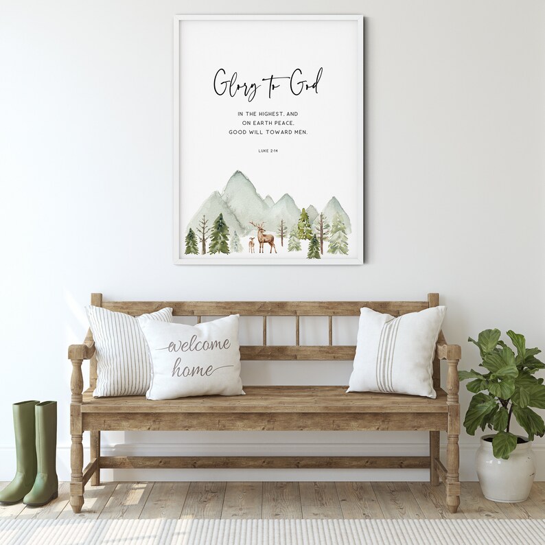 May include: A watercolor print with a mountain scene and a deer and fawn. The print has the text "Glory to God" and a Bible verse from Luke 2:14. The print is framed and hanging on a white wall above a wooden bench.