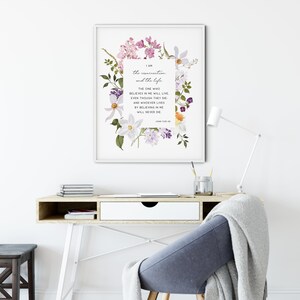 May include: A framed print with a white background and a floral border. The text in the centre reads "I am the resurrection and the life. The one who believes in me will live, even though they die. And whoever lives by believing in me will never die. John 11:25-26".