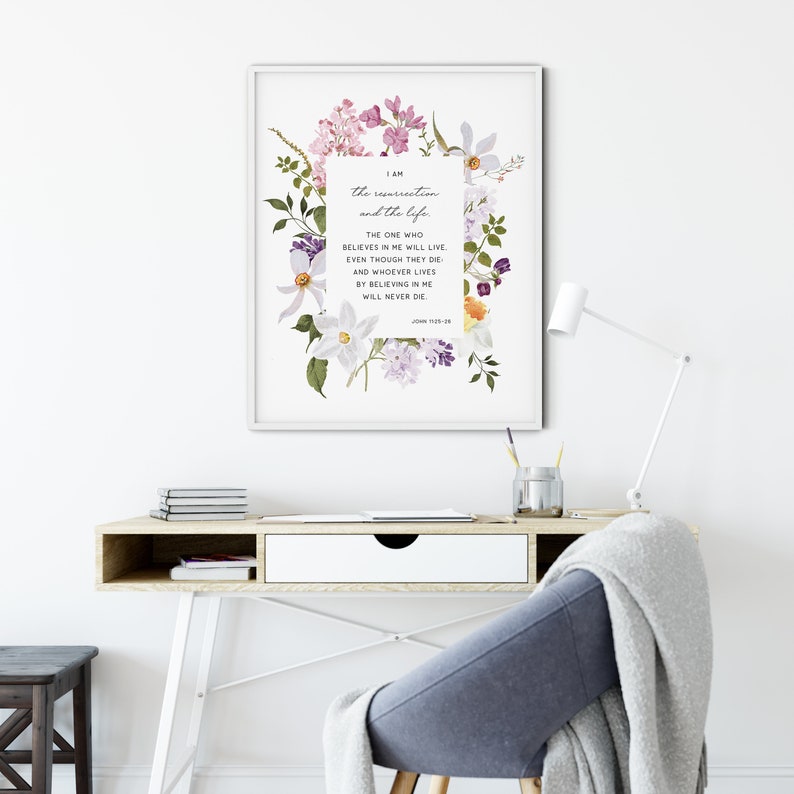 May include: A framed print with a white background and a floral border. The text in the centre reads "I am the resurrection and the life. The one who believes in me will live, even though they die. And whoever lives by believing in me will never die. John 11:25-26".