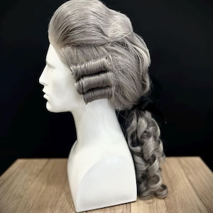 May include: A silver-gray wig styled in a historical fashion with three horizontal barrel curls on each side of the head, a voluminous top section, and a long, curled ponytail secured at the nape of the neck with a black ribbon on a mannequin.