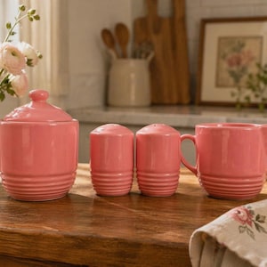 May include: A pink ceramic kitchen set including a lidded sugar bowl, two salt and pepper shakers, and a creamer with a handle. The set has a ribbed design and is displayed on a wooden surface.