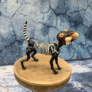 May include: A black and white skeletal dog figurine with a large, toothy, red mouth. The figurine is standing on a small wooden table with a round top. The table is surrounded by brown branches.