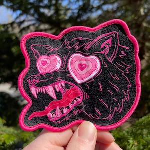 May include: Embroidered patch featuring a black wolf with pink heart-shaped eyes and a lolling tongue. The patch has a pink border and is likely designed for clothing or accessories. The design is bold and eye-catching.