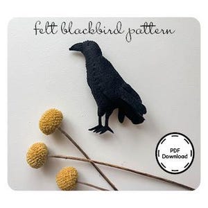 May include: A black felt blackbird stands next to three yellow billy buttons on a white background. The text "felt blackbird pattern" is above the bird. A PDF download icon is in the lower right corner.