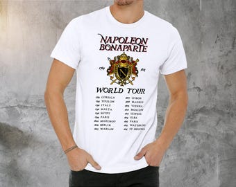Könnte beinhalten: Weißes T-Shirt mit einem Grafikdruck des Wappens von Napoleon Bonaparte und einer Liste der Städte, die er auf seiner Welttournee besucht hat. Der Text lautet "Napoleon Bonaparte 1769 Welttournee 1815" und listet Städte wie Korsika, Toulon, Italien, Ägypten, Lissabon, Madrid, Wien, Moskau, Leipzig, Malta, Paris, Elba, Marengo, Paris, Berlin, Waterloo, Warschau und St. Helena auf.