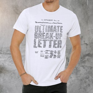 May include: White t-shirt with the text "ULTIMATE BREAK-UP LETTER" in a black, newspaper-style font. The shirt also features text at the top that reads "In Congress, July 4, 1776". The shirt is short-sleeved.