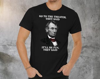 May include: Black t-shirt featuring a portrait of Abraham Lincoln with a red bow tie. The shirt includes the text "GO TO THE THEATER, THEY SAID" above the portrait and "IT'LL BE FUN, THEY SAID" below.