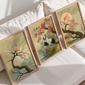 May include: Three rectangular framed wall art prints featuring nature scenes. The left print shows a bare tree with pink blossoms. The center print depicts a crane standing near pink flowers. The right print displays a full moon behind a flowering tree.