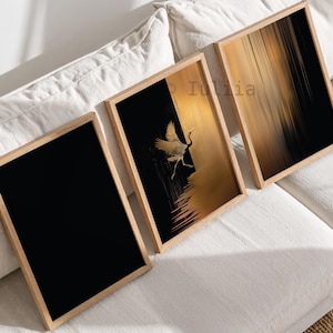 May include: Three rectangular wall art prints in light wood frames. The left print is solid black. The center print features a white crane in flight against a black and gold background. The right print displays a vertical gold and black abstract pattern.