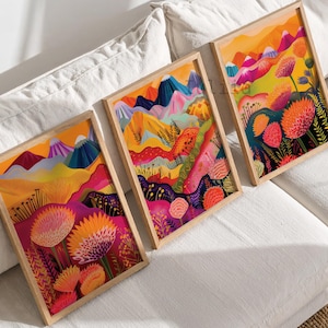 Colorful 3 piece wall art displayed in natural wood frames on soft neutral bedding. The vibrant triptych features abstract mountains, bold orange and pink flowers, layered landscape shapes and rich yellow, magenta and blue tones, creating playful maximalist wall decor for bedrooms, living rooms and colorful contemporary interiors.