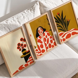 May include: Three framed rectangular wall art prints featuring illustrations with a color palette of mustard yellow, cream, black, and bright orange. The designs include a person with black hair, a potted plant, and floral motifs with organic shapes.