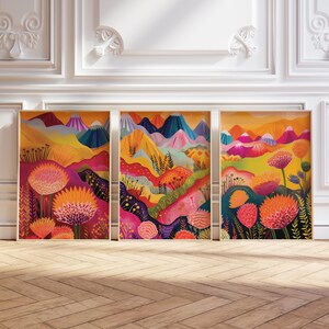 Funky floral triptych displayed in three framed panels against an elegant light wall with decorative molding. The artwork combines bright orange, pink, yellow and deep blue landscape shapes with oversized botanical forms and abstract mountains, creating colorful statement wall art for maximalist, eclectic and dopamine-inspired interiors.