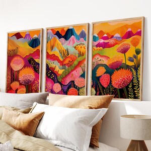 Colorful wall art set of 3 displayed above a neutral bed in a warm modern bedroom. The bold prints feature abstract mountain scenery, vibrant pink and orange florals, yellow landscape layers and deep blue accents, creating energetic botanical wall decor and a playful focal point for bright contemporary spaces.