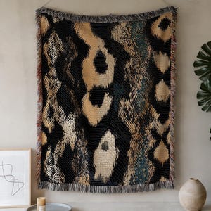 May include: A woven throw blanket with a snakeskin pattern in black, tan, and teal. The blanket has a fringed edge and is hanging on a wall. The design features large, irregular shapes resembling reptile scales.
