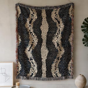 May include: A patterned throw blanket with a design of black and cream-coloured dots and vertical stripes. The blanket has a fringed edge and is hanging on a wall. The colour scheme includes black, cream, and blue.