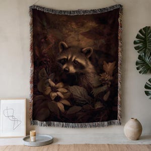 May include: A woven throw blanket with a detailed illustration of a raccoon amongst leaves and flowers. The artwork uses warm, earthy tones of brown, beige, and sepia, with a fringed edge. The blanket is designed as a decorative home accent.