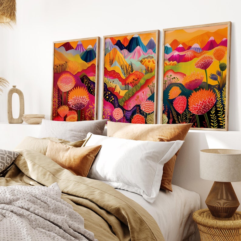 Colorful wall art set of 3 displayed above a neutral bed in a warm modern bedroom. The bold prints feature abstract mountain scenery, vibrant pink and orange florals, yellow landscape layers and deep blue accents, creating energetic botanical wall decor and a playful focal point for bright contemporary spaces.