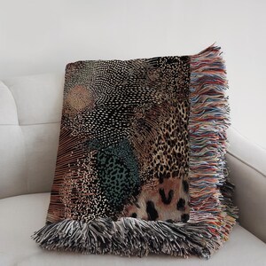 Op de afbeelding: Een geweven plaid met een complex patroon van zwarte, beige en blauwgroene stippen, lijnen en luipaardprintmotieven. De deken heeft een gestructureerd oppervlak en is afgewerkt met lange, veelkleurige franjes langs de zij- en onderranden.