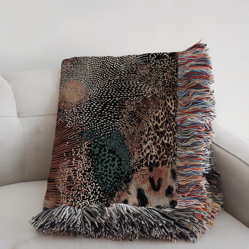 Op de afbeelding: Een geweven plaid met een complex patroon van zwarte, beige en blauwgroene stippen, lijnen en luipaardprintmotieven. De deken heeft een gestructureerd oppervlak en is afgewerkt met lange, veelkleurige franjes langs de zij- en onderranden.