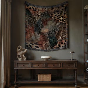 Op de afbeelding: Een rechthoekig geweven wandtapijt met een complex, meerkleurig dierenprintpatroon in tinten bruin, zwart, beige en groen. De stof heeft een getextureerd oppervlak en is afgewerkt met een gefranjerde rand langs de onderkant.