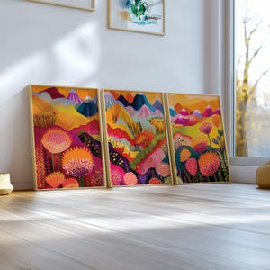 Colorful 3 piece wall art leaning on a bright white windowsill in natural daylight. The coordinated prints feature abstract floral landscapes with orange, pink, yellow, green and deep blue tones, combining playful botanical forms and mountain shapes in a vibrant modern triptych.