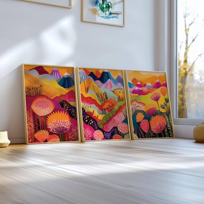 Colorful 3 piece wall art leaning on a bright white windowsill in natural daylight. The coordinated prints feature abstract floral landscapes with orange, pink, yellow, green and deep blue tones, combining playful botanical forms and mountain shapes in a vibrant modern triptych.