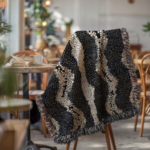 Peut inclure: Une couverture &agrave; motifs noirs et beiges drap&eacute;e sur une chaise en bois. La couverture pr&eacute;sente un motif de points r&eacute;p&eacute;t&eacute;s avec des rayures beiges verticales. Les bords sont finis avec une frange. La couverture est dans un cadre de caf&eacute;.