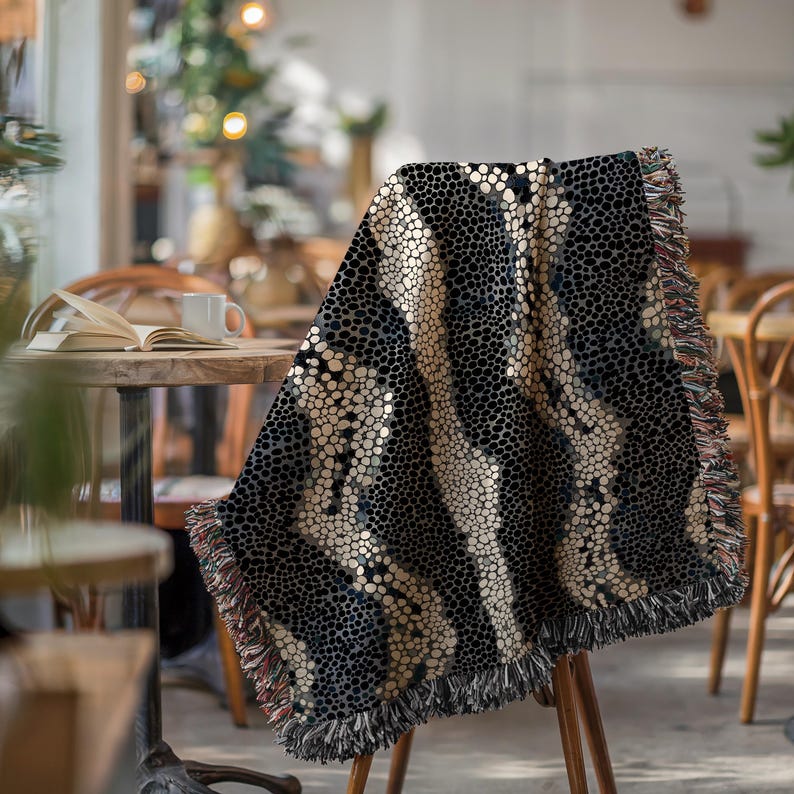 Peut inclure: Une couverture &agrave; motifs noirs et beiges drap&eacute;e sur une chaise en bois. La couverture pr&eacute;sente un motif de points r&eacute;p&eacute;t&eacute;s avec des rayures beiges verticales. Les bords sont finis avec une frange. La couverture est dans un cadre de caf&eacute;.