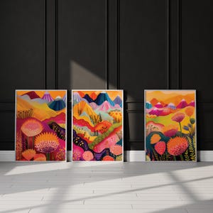 Three bold color prints leaning against a dark charcoal wall in strong natural sunlight. The vibrant triptych features abstract mountains, layered landscapes and oversized floral forms in orange, pink, yellow and deep blue, creating dramatic colorful wall art for eclectic, maximalist and contemporary interiors.