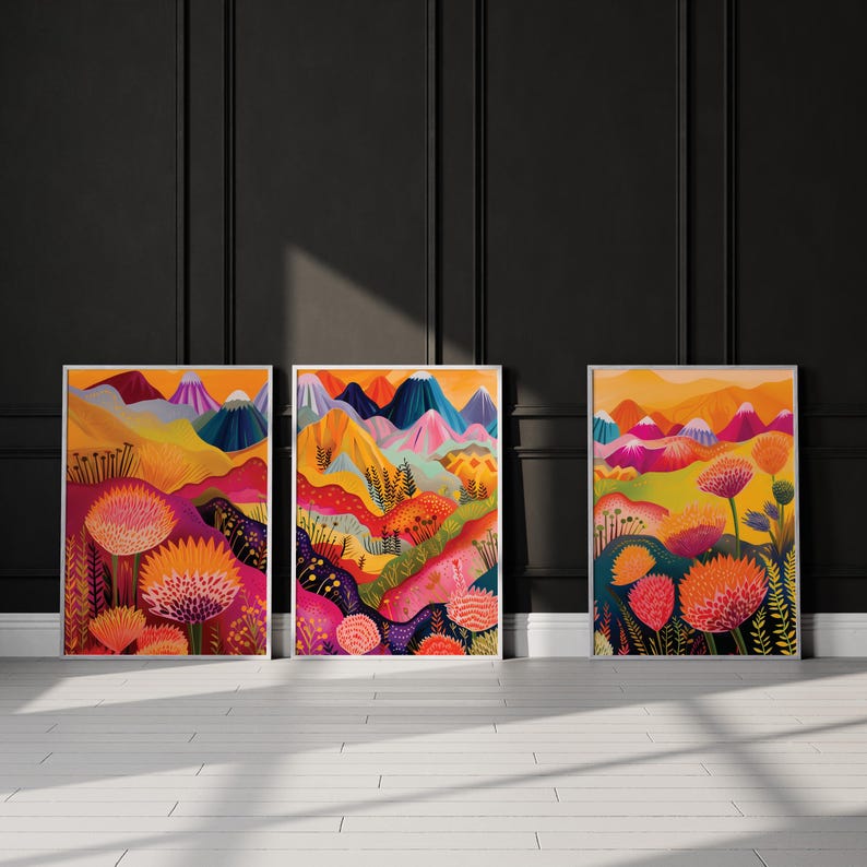 Three bold color prints leaning against a dark charcoal wall in strong natural sunlight. The vibrant triptych features abstract mountains, layered landscapes and oversized floral forms in orange, pink, yellow and deep blue, creating dramatic colorful wall art for eclectic, maximalist and contemporary interiors.
