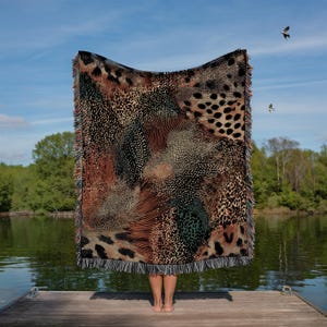 Op de afbeelding: Een rechthoekige geweven plaid met een complex, meerkleurig dierenprintpatroon met luipaardvlekken, jachtluipaardstippen en abstracte texturen in tinten zwart, bruin, beige en groen, afgewerkt met een franjerand aan alle zijden.