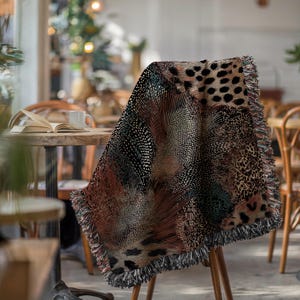 Op de afbeelding: Een geweven plaid met een veelkleurig abstract patroon met luipaardvlekken, fijne lijnen en gestippelde texturen. De deken heeft een rand met franjes met veelkleurige draden en toont een mix van zwarte, bruine en beige tinten.