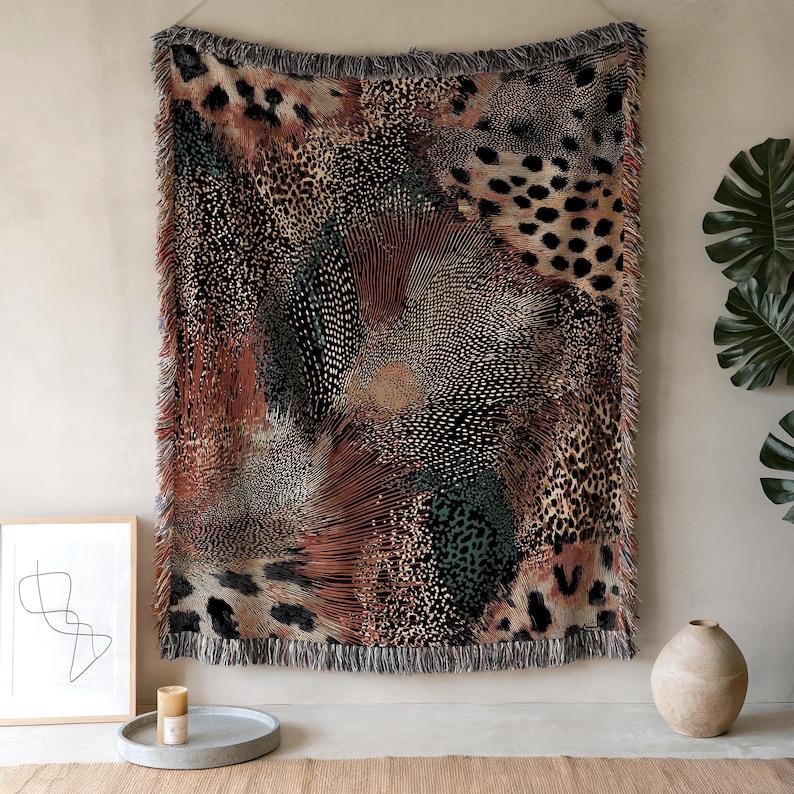 Op de afbeelding: Een rechthoekig geweven wandtapijt met een complex abstract patroon met dierenprints, waaronder luipaardvlekken en tijgerstrepen in tinten zwart, bruin, roest en gedempt groen, afgewerkt met een decoratieve rand met franjes.